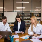 Three colleagues in a modern office setting are working together. They're seated around a table with a laptop, papers, and open books, appearing engaged and collaborative.