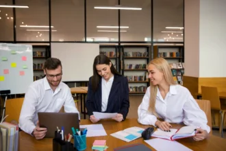 Three colleagues in a modern office setting are working together. They're seated around a table with a laptop, papers, and open books, appearing engaged and collaborative.
